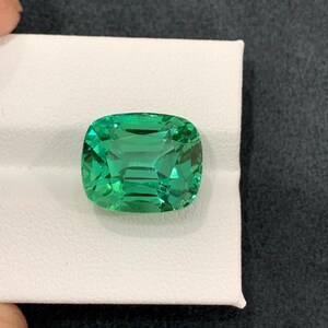 May include: A vibrant, cushion-cut emerald gemstone. The gem is a vivid green color and sits on a white surface. The gemstone's facets reflect light, creating a sparkling effect. The gem is a square shape with rounded corners.