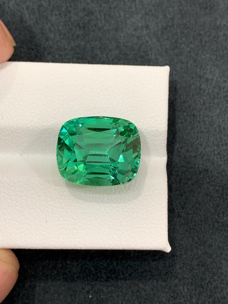 May include: A vibrant, cushion-cut emerald gemstone. The gem is a vivid green color and sits on a white surface. The gemstone's facets reflect light, creating a sparkling effect. The gem is a square shape with rounded corners.