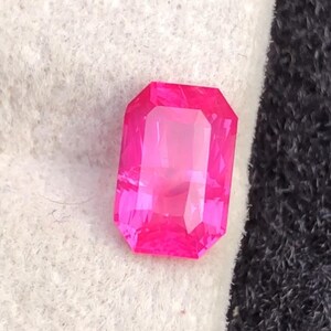 May include: A vibrant, rectangular-cut, hot pink gemstone. The gem has a faceted surface that reflects light, creating a brilliant shine. The stone is set against a white and black background.