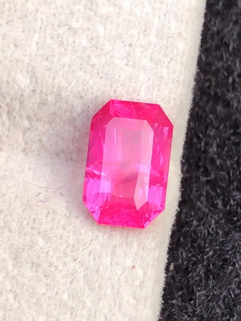 May include: A vibrant, rectangular-cut, hot pink gemstone. The gem has a faceted surface that reflects light, creating a brilliant shine. The stone is set against a white and black background.