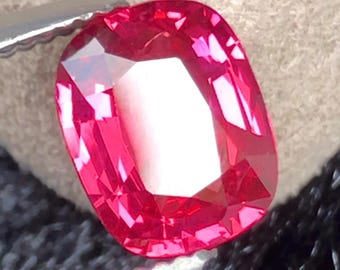 Puede incluir: Una gema roja vibrante de corte cojín con una superficie facetada que refleja la luz. La piedra está sujeta por una herramienta metálica, mostrando su profundo color carmesí, alta claridad y un corte geométrico simétrico con reflejos.