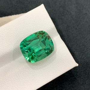 May include: A vibrant, emerald-green gemstone with a cushion cut. The gem is displayed on a white surface, showcasing its facets and clarity. The stone's color is a rich, saturated green, reflecting light.