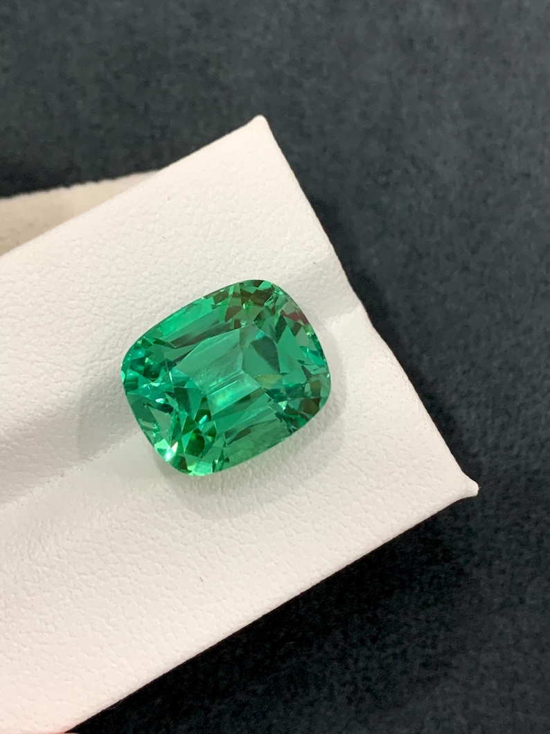 May include: A vibrant, emerald-green gemstone with a cushion cut. The gem is displayed on a white surface, showcasing its facets and clarity. The stone's color is a rich, saturated green, reflecting light.