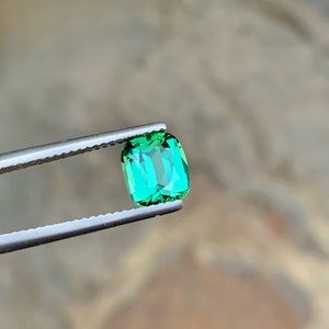 May include: A vibrant green gemstone with a rectangular cut, held by tweezers against a blurred background.