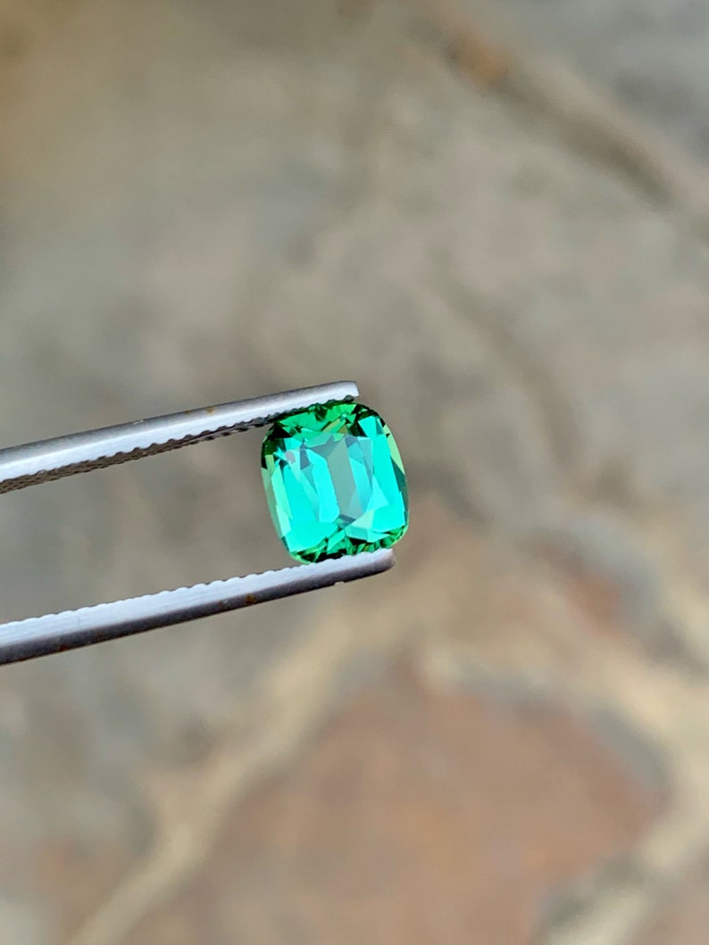 May include: A vibrant green gemstone with a rectangular cut, held by tweezers against a blurred background.