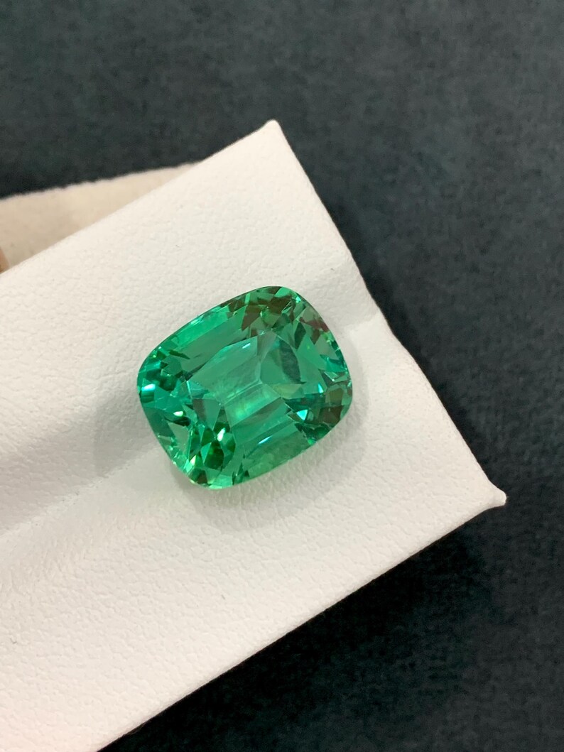 May include: A vibrant, emerald-green gemstone with a cushion cut. The gem is set against a white surface, showcasing its clarity and brilliance. The facets reflect light, creating a sparkling effect. The gemstone's shape is rectangular with rounded corners.