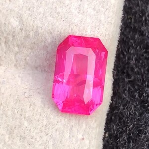 May include: A vibrant, rectangular-cut, hot pink gemstone. The gem has a faceted surface that reflects light, creating a sparkling effect. The gemstone is set against a white and black background.