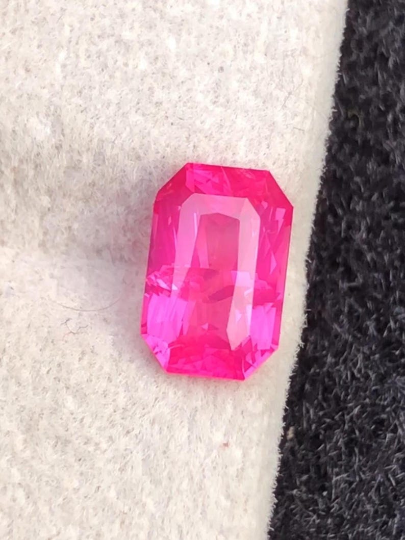 May include: A vibrant, rectangular-cut, hot pink gemstone. The gem has a faceted surface that reflects light, creating a sparkling effect. The gemstone is set against a white and black background.