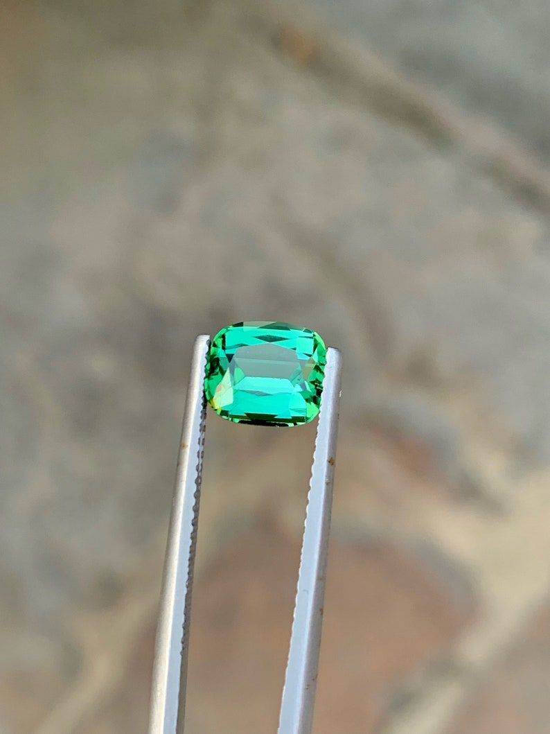 May include: A vibrant green gemstone, likely an emerald, is held in tweezers. The gemstone is rectangular in shape with a slight emerald cut.