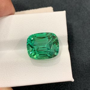 May include: A vibrant, emerald-cut green gemstone with a cushion shape. The gem is held on a white surface, showcasing its facets and clarity. The stone's color is a rich, saturated green, reflecting light.