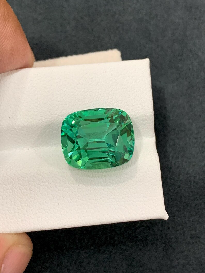 May include: A vibrant, emerald-cut green gemstone with a cushion shape. The gem is held on a white surface, showcasing its facets and clarity. The stone's color is a rich, saturated green, reflecting light.