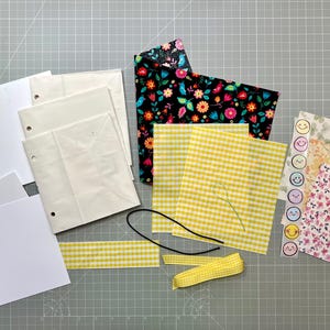 May include: A collection of craft supplies including white paper, patterned fabric, yellow gingham fabric, a black cord, yellow ribbon, and stickers with smiley faces. The fabric features floral and gingham patterns.