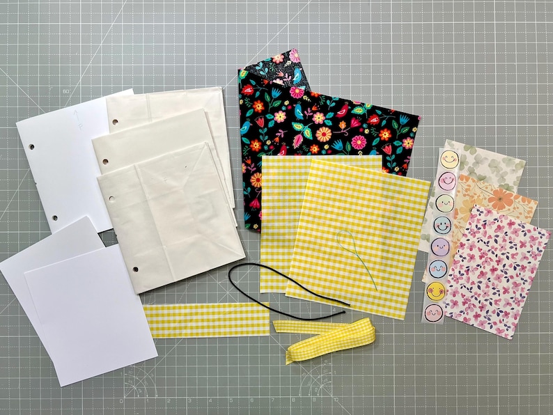 May include: A collection of craft supplies including white paper, patterned fabric, yellow gingham fabric, a black cord, yellow ribbon, and stickers with smiley faces. The fabric features floral and gingham patterns.