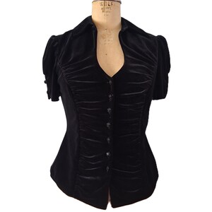 May include: A black velvet blouse featuring short puff sleeves, a collared neckline, and a front button closure. The bodice is detailed with vertical ruching along the center panel, and the sleeves are finished with small decorative buttons.