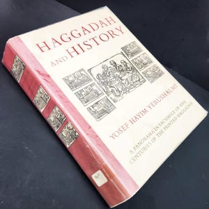 May include: A hardcover book titled "Haggadah and History" with red and cream colors. The book features illustrations and text, including the author's name, Yosef Hayim Yerushalmi. The book is wrapped in clear plastic.