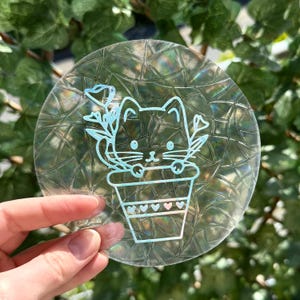 Puede incluir: Una pegatina redonda transparente con un diseño de gato en una maceta. El gato y la maceta están delineados en azul claro. La pegatina tiene una superficie texturizada e iridiscente, y la sostiene una mano.