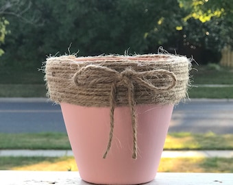 Könnte beinhalten: Ein rosa bemalter Blumentopf aus Terrakotta mit dekorativem Juteseilbesatz. Das Seil ist um den oberen Teil des Topfes gewickelt und mit einer Schleife vorne gebunden. Der Topf steht auf einer weißen Oberfläche.