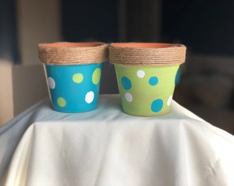 Könnte beinhalten: Zwei kleine Terrakotta-Töpfe mit einer Jute-Verzierung. Ein Topf ist in Türkisblau mit weißen und grünen Punkten bemalt. Der andere Topf ist in Grün mit weißen und blauen Punkten bemalt.