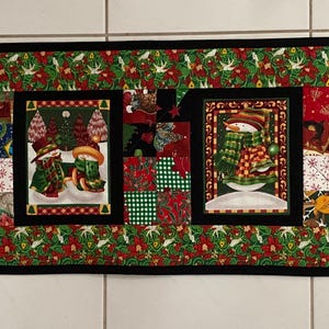 May include: A rectangular Christmas table runner with a black border and a festive holly and berry pattern. The runner features five rectangular panels with holiday-themed images, including snowmen, Christmas trees, and a witch.