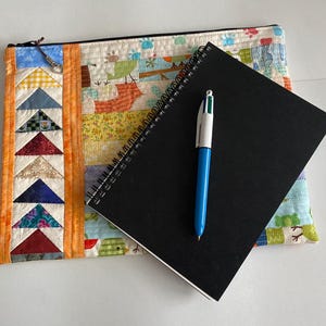 May include: A black spiral-bound notebook and a blue pen rest on a colourful quilted pouch. The pouch features a patchwork design with various fabrics and a zip closure. The notebook is positioned at an angle.