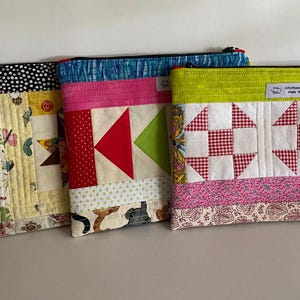 May include: Three handmade quilted zipper pouches. One has a black and white polka dot top, another a red and green geometric design, and the third a red and white checkered pattern. Each pouch features a unique fabric design. The pouches are in various colors.