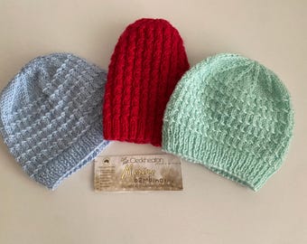 May include: Three knitted baby hats in different colours: light blue, red and light green. The hats have a textured pattern and are made with soft yarn. The hats are arranged on a white surface with a small tag that reads "Clekheatons Merino Bambinosy".