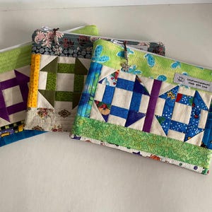 May include: Three handmade quilted zipper pouches with patchwork designs. The pouches feature various colours, including blue, green, purple, and yellow, with floral and geometric patterns. Each pouch has a different charm on the zipper.