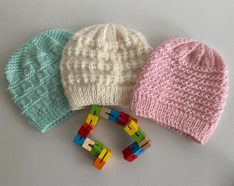 May include: Three knitted hats in pastel shades: pale blue, cream, and pink. The hats feature various textured patterns. A colourful wooden puzzle is placed in front of the hats.