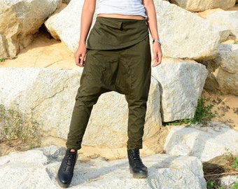 May include: Olive green drop-crotch pants featuring a wide, folded waistband with a flap detail and tapered legs that gather at the ankles. The pants are paired with a plain white t-shirt and black lace-up boots, showcasing a relaxed fit.