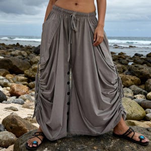 Peut inclure: Pantalon sarouel gris avec taille élastique à cordon, poches latérales et fermeture boutonnée. Le pantalon présente des détails froncés sur les côtés et est porté avec des sandales noires à détails turquoise. Tissu souple.