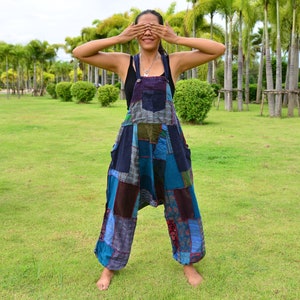 Może przedstawiać: Kobieta w kombinezonie patchwork w odcieniach niebieskiego, zielonego i fioletowego. Stoi na trawiastym terenie z palmami w tle.