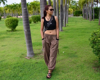 May include: A woman wearing a black crop top with the word "Free" on it, brown pants, and a brown leather strap bag. She is standing in a park with palm trees in the background.