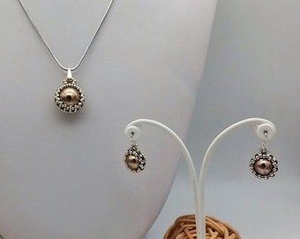 May include: A jewelry set featuring a necklace and earrings. The necklace has a silver chain with a pendant showcasing a large, brown pearl surrounded by small, silver beads. The earrings mirror the pendant design. The set is displayed on a white mannequin.