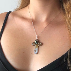 Può includere: Una collana d'argento con un ciondolo a forma di croce. La croce presenta un mix di colori, tra cui bianco, oro e verde. La collana è indossata su un top nero.