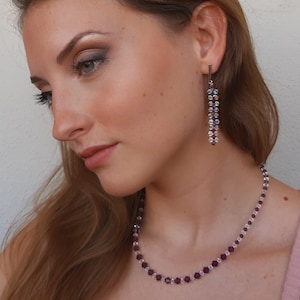 Può includere: Una donna indossa una collana e orecchini. La collana e gli orecchini sono fatti di piccole perline sfaccettate viola e trasparenti. Gli orecchini pendono in linea verticale. La donna indossa un top rosa.