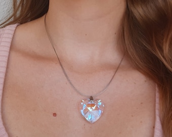 Pode incluir: Um colar com um pingente transparente em forma de coração e uma corrente prateada. O pingente é facetado, refletindo a luz com um efeito arco-íris. O colar é usado.