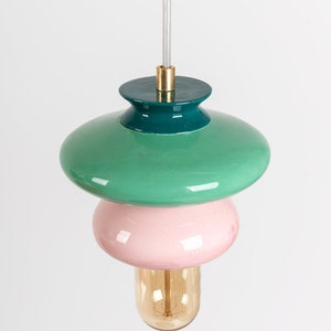 May include: A modern pendant light with three tiers. The top tier is a green saucer, the middle tier is a pink sphere, and the bottom tier is a clear glass bulb.