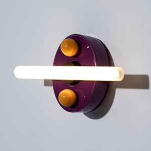 May include: A modern wall sconce with a purple circular base, two yellow spherical accents, and a horizontal, illuminated white tube light. The fixture is mounted on a white wall.
