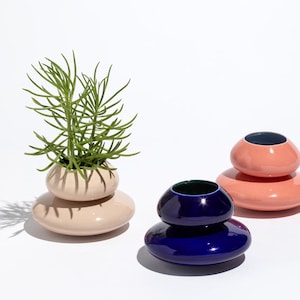 May include: Three ceramic decorative vases in shades of pink and blue. One vase holds a green plant. The vases have a stacked, pebble-like design. The background is white.