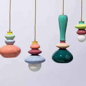 Op de afbeelding: Vier kleurrijke hanglampen met gestapelde, afgeronde vormen in tinten koraal, turquoise, roze en blauw. Elke lamp hangt aan een bruin snoer met een goudkleurige armatuur aan de bovenkant. De lampen hebben een modern, artistiek ontwerp.