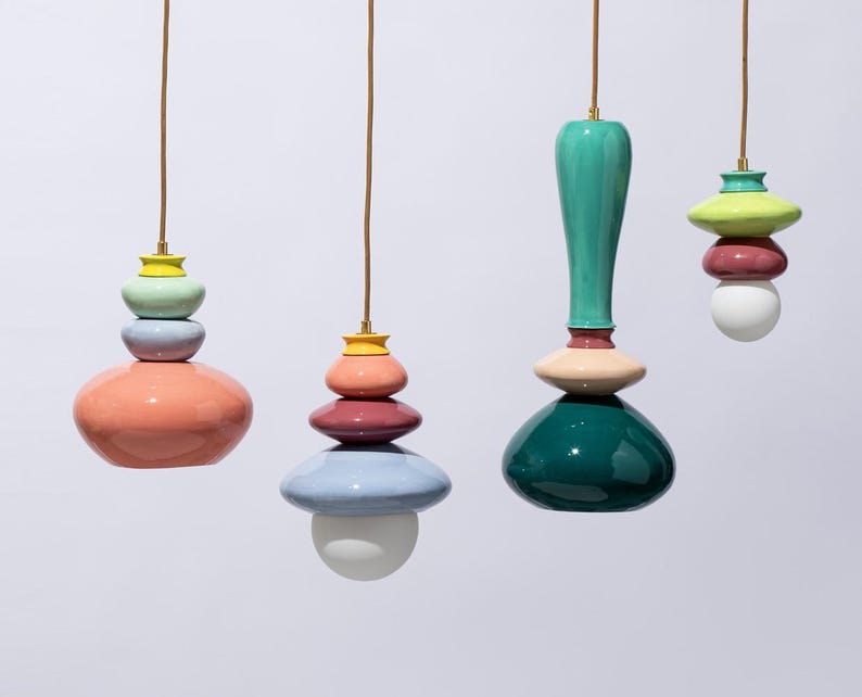 Op de afbeelding: Vier kleurrijke hanglampen met gestapelde, afgeronde vormen in tinten koraal, turquoise, roze en blauw. Elke lamp hangt aan een bruin snoer met een goudkleurige armatuur aan de bovenkant. De lampen hebben een modern, artistiek ontwerp.