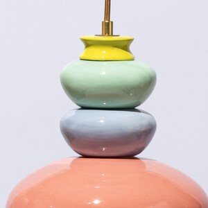 Op de afbeelding: Een veelkleurige, gestapelde keramische hanglamp. De lamp heeft een koraalkleurige basis, een lichtblauw middengedeelte, een mintgroen midden en een gele bovenkant. De lamp wordt opgehangen aan een goudkleurig snoer.