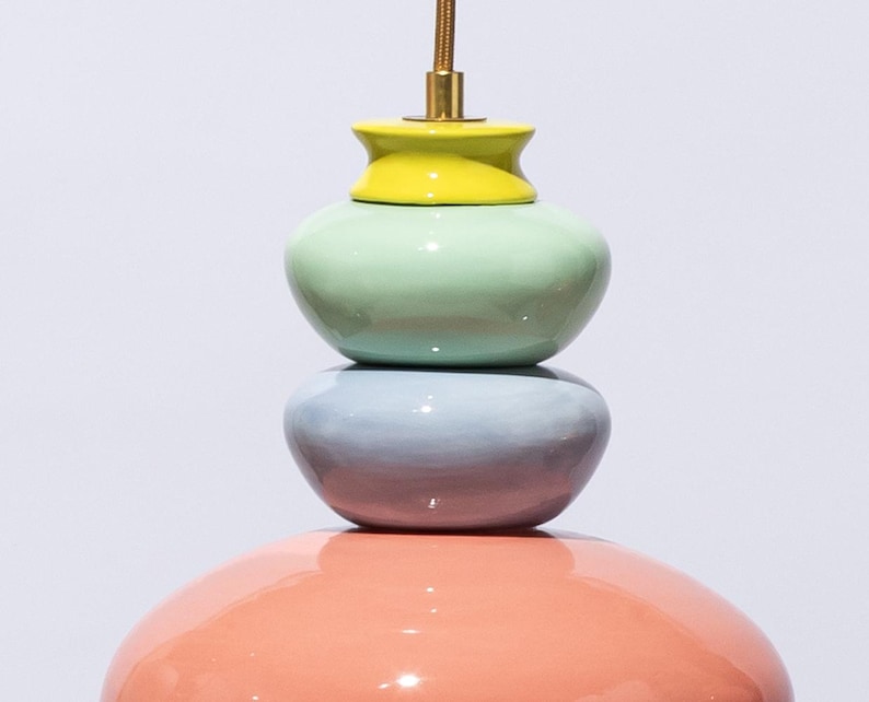 Op de afbeelding: Een veelkleurige, gestapelde keramische hanglamp. De lamp heeft een koraalkleurige basis, een lichtblauw middengedeelte, een mintgroen midden en een gele bovenkant. De lamp wordt opgehangen aan een goudkleurig snoer.