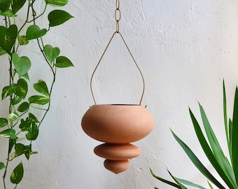 Puede incluir: Una maceta colgante de terracota con un diseño único y en capas. La maceta está suspendida por una fina cadena de metal de color dorado. La forma redonda y apilada añade un toque moderno. Las plantas verdes rodean la maceta, creando una estética natural.