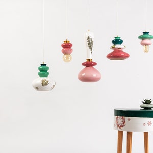 May include: Five unique pendant lights with colorful ceramic shades and gold accents hang from the ceiling. The lights are suspended over a small, round, white table with green and pink floral designs and wooden legs.
