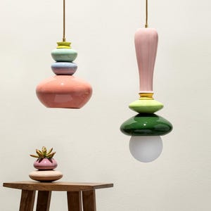 Op de afbeelding: Twee kleurrijke, gestapelde keramische hanglampen hangen aan het plafond. E&eacute;n heeft een roze basis, met blauwe, gele en groene accenten. De andere heeft een roze bovenkant, groene en gele accenten en een witte bol. Een kleine vetplant staat op een houten tafel.