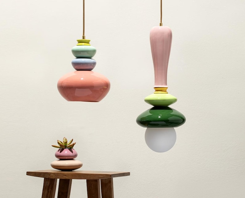 Op de afbeelding: Twee kleurrijke, gestapelde keramische hanglampen hangen aan het plafond. E&eacute;n heeft een roze basis, met blauwe, gele en groene accenten. De andere heeft een roze bovenkant, groene en gele accenten en een witte bol. Een kleine vetplant staat op een houten tafel.