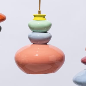 Op de afbeelding: Kleurrijke, gestapelde keramische hanglampen in tinten koraal, groen, blauw en geel. Elke lamp heeft een unieke vorm en is opgehangen aan een goudkleurig snoer. De achtergrond is wit.
