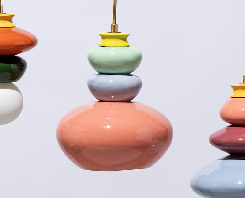 Op de afbeelding: Kleurrijke, gestapelde keramische hanglampen in tinten koraal, groen, blauw en geel. Elke lamp heeft een unieke vorm en is opgehangen aan een goudkleurig snoer. De achtergrond is wit.