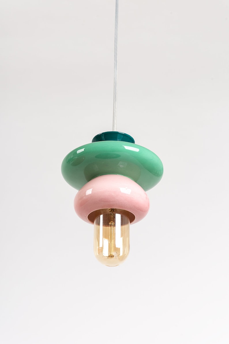 May include: A modern pendant light with three stacked ceramic shades in green, pink, and teal. The light has a clear glass bulb.